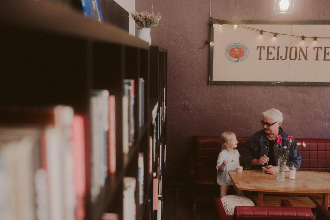 Isoäiti ja lapsi jäätelöllä Cafe Mathildedalin violeteilla sohvilla, vasemmalla sivulla kirjahylly.